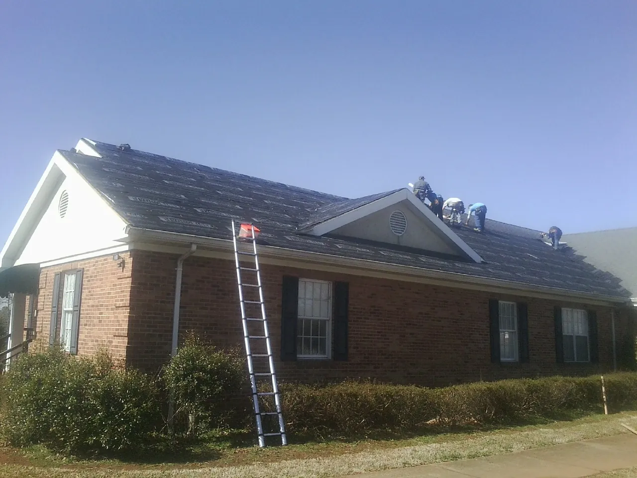 Crew tearing shingles off a brick ranch roof with a ladder set — Emergency Roof Repair work by our crew in Lawton